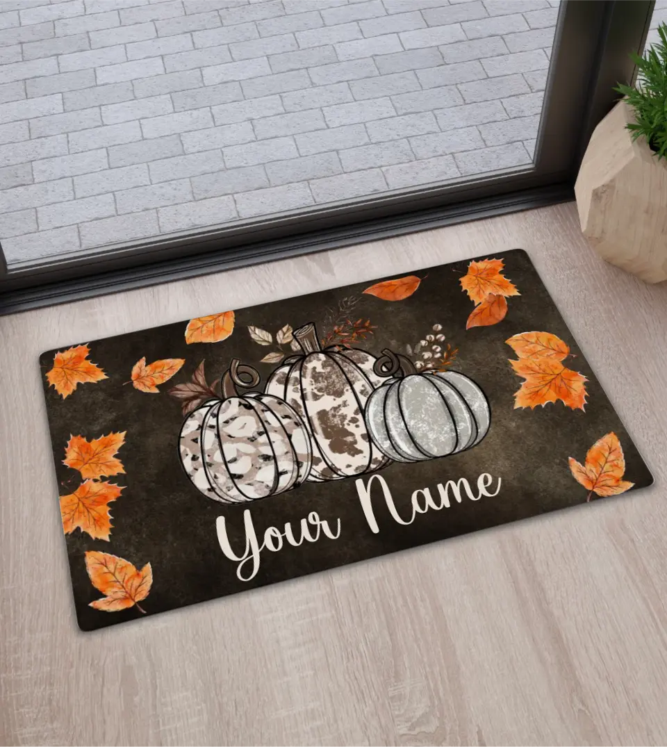 A rectangular door mat displays a3cowhide pumpkins. Below the pumpkins is a customizable family name. Surrounding the pumpkin are subtle fall leaves, creating a welcoming fall ambiance. Ideal for autumn decor or as a personalized Halloween gift.