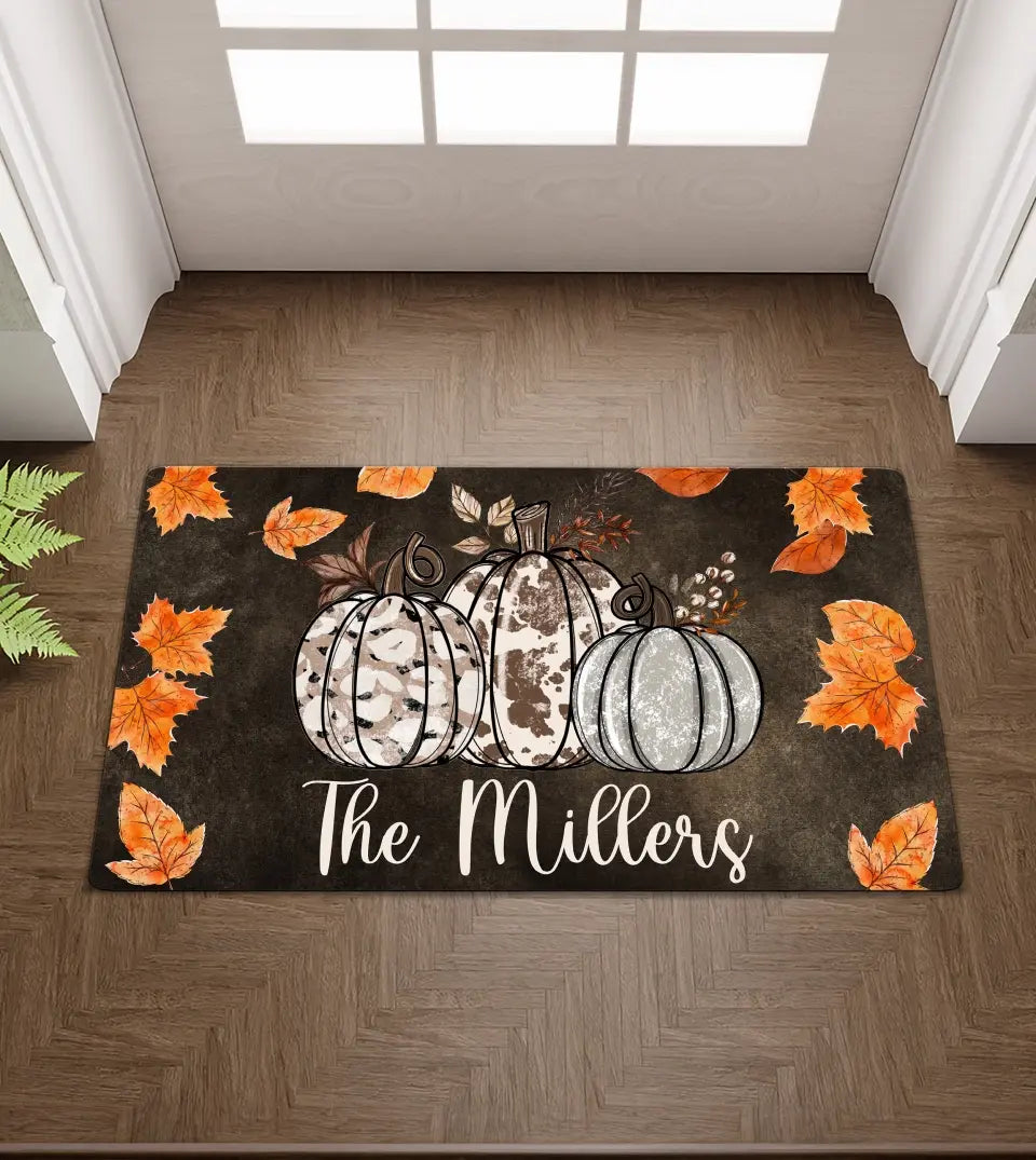 A rectangular door mat displays a3cowhide pumpkins. Below the pumpkins is a customizable family name. Surrounding the pumpkin are subtle fall leaves, creating a welcoming fall ambiance. Ideal for autumn decor or as a personalized Halloween gift.