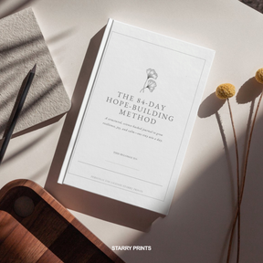 Book titled 'The 84-Day Hope-Building Method' by Starry Prints on a light surface with a pen and dried flowers.